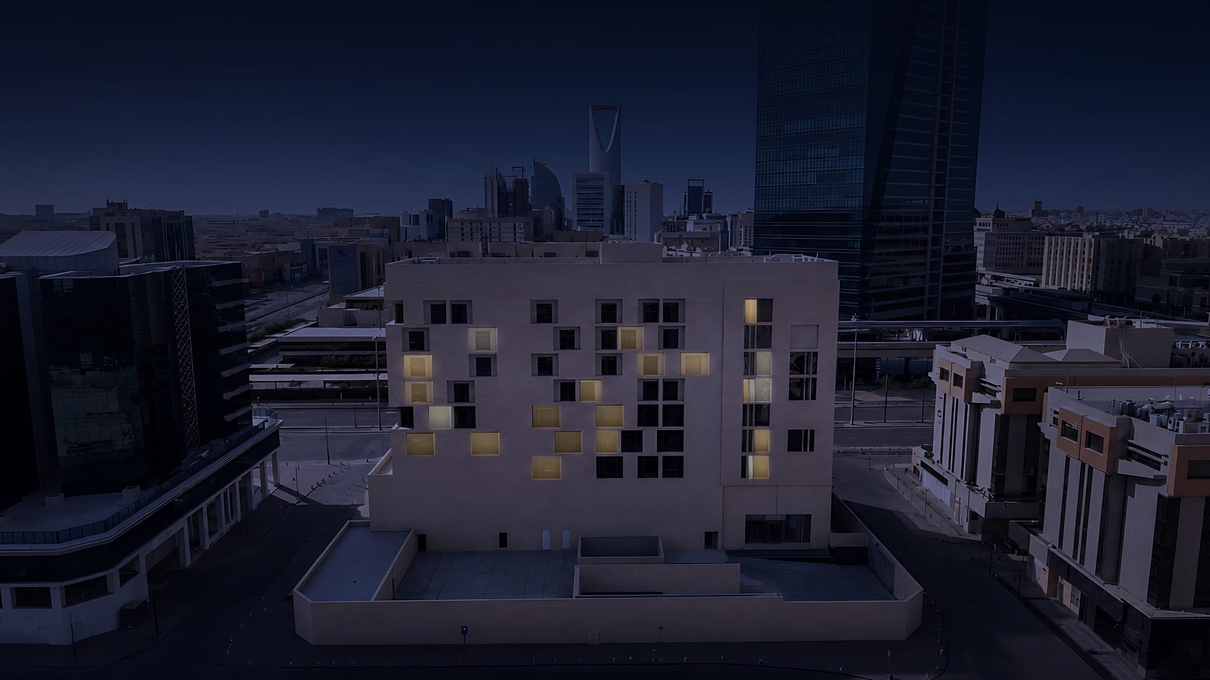 Aerial photograph from behind the building at nightfall, rear windows lit, looking toward the Riyadh skyline with Kingdom Centre on the horizon.