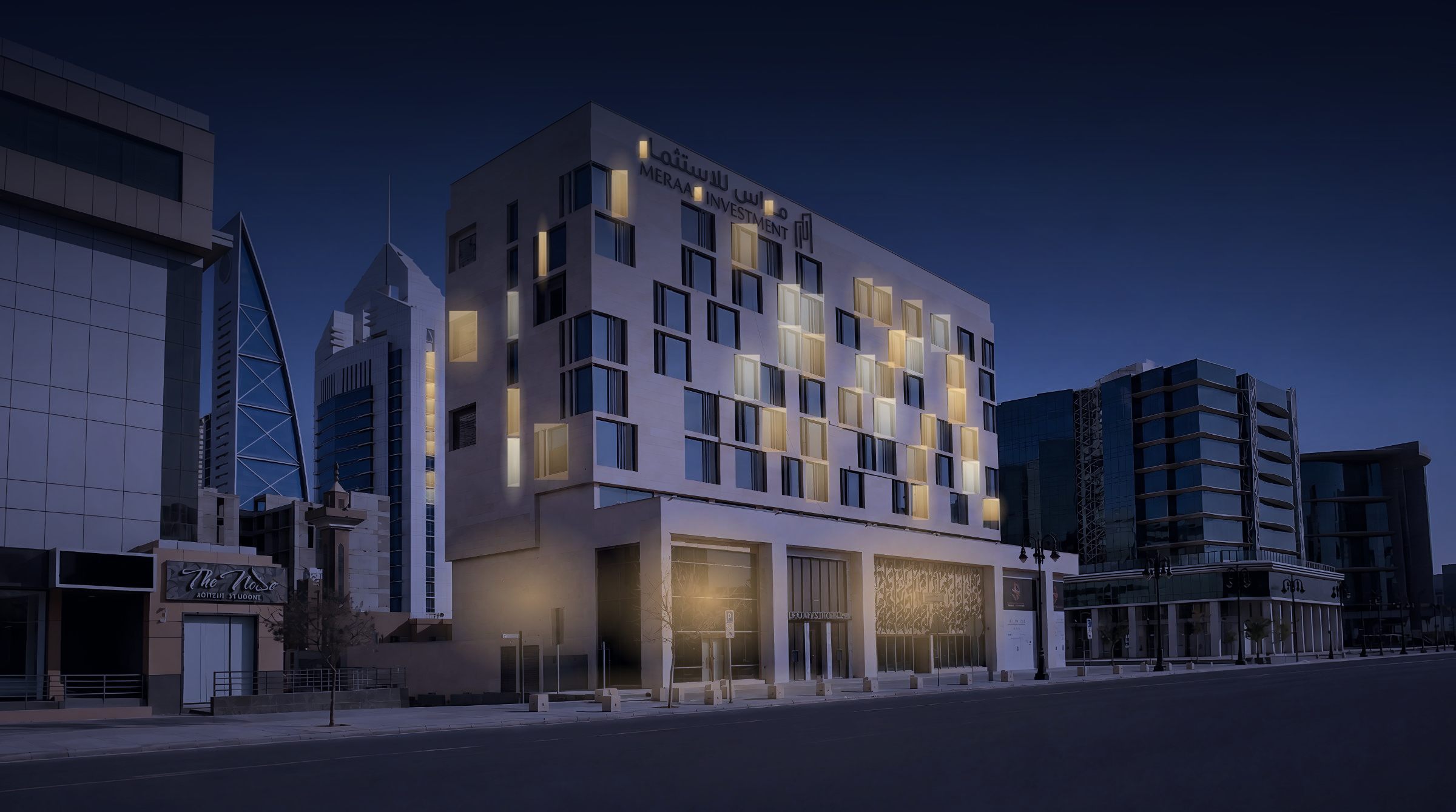 The SBS Business Tower street facade at night: lit windows scattered across the beige stone grid, the retail podium glowing, the Al-Olaya towers rising behind.