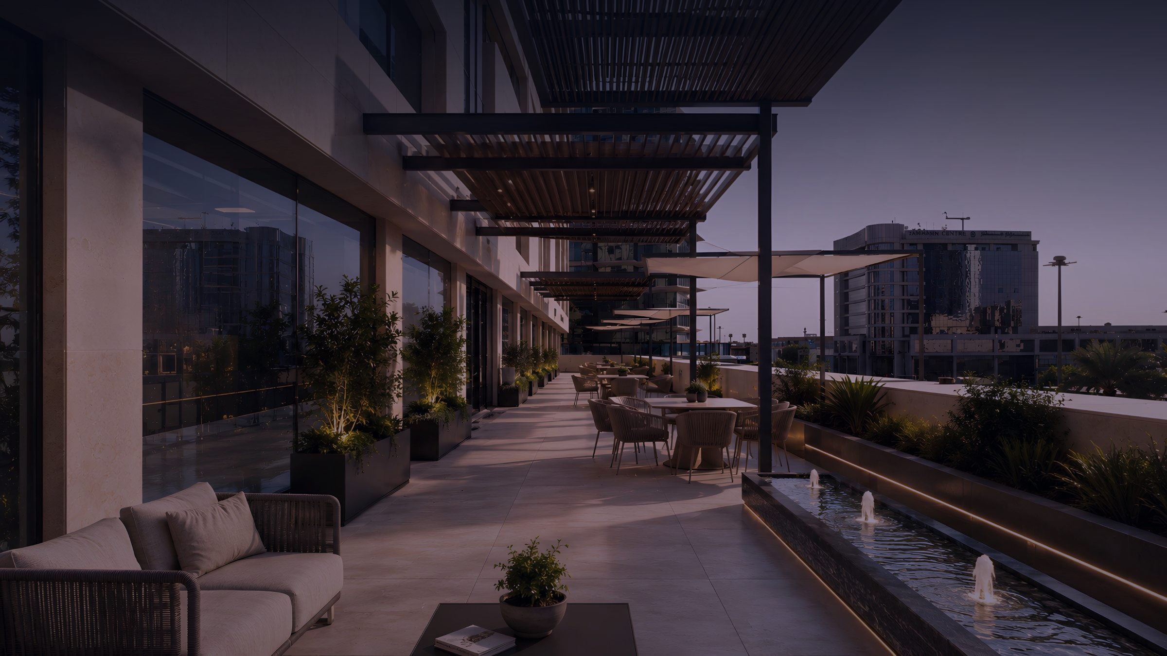 The office terrace at dusk: timber pergolas, planter uplights, cafe seating and a lit water feature overlooking Al-Olaya.